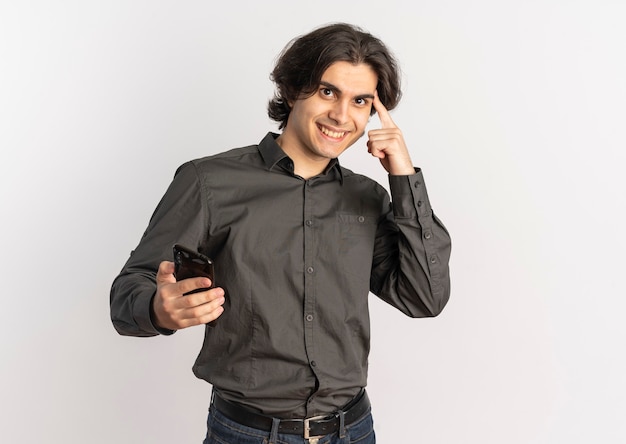 Il giovane uomo caucasico bello sorridente tiene il telefono e mette il dito sulla testa isolata su fondo bianco con lo spazio della copia