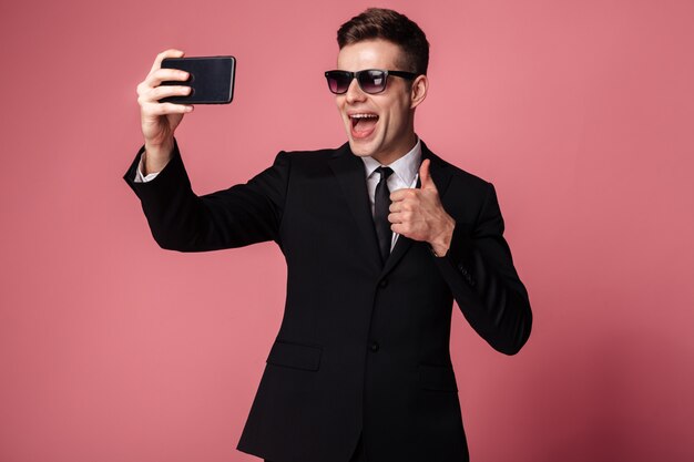 Il giovane uomo d'affari allegro fa il selfie con i pollici in su dal telefono.