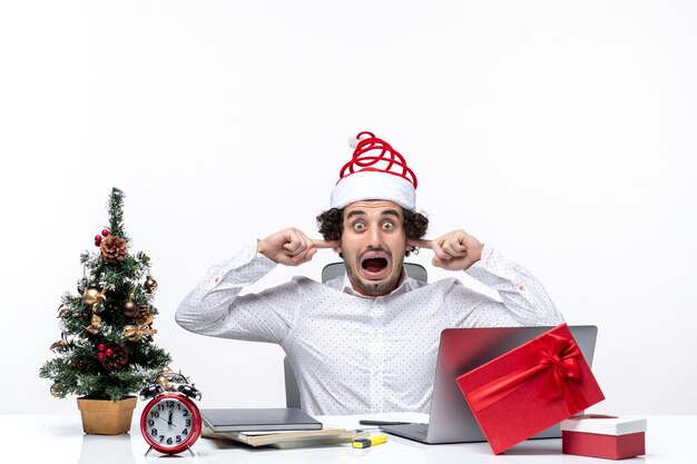 Il giovane uomo d'affari arrabbiato con il cappello di Babbo Natale divertente che celebra il Natale chiudendo le sue orecchie impedisce l'ascolto in ufficio su priorità bassa bianca