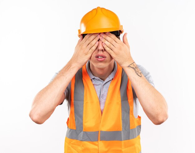 Il giovane uomo del costruttore in uniforme ha coperto gli occhi di mani isolate sulla parete bianca