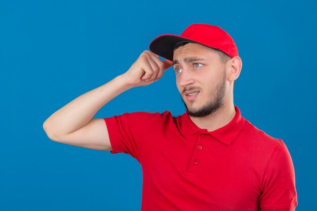 Il giovane uomo di consegna che indossa la camicia di polo rossa e il cappuccio confuso si sente dubbioso e insicuro su sfondo blu isolato