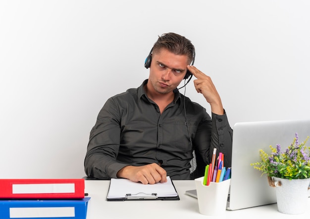 Il giovane uomo di lavoratore di ufficio biondo infastidito sulle cuffie si siede alla scrivania con strumenti di ufficio utilizzando gli occhi rotanti del computer portatile isolati su priorità bassa bianca con lo spazio della copia