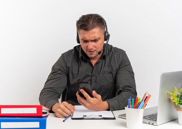 Il giovane uomo di lavoratore di ufficio biondo infastidito sulle cuffie si siede alla scrivania con strumenti di ufficio utilizzando il computer portatile esamina il telefono isolato su priorità bassa bianca con lo spazio della copia