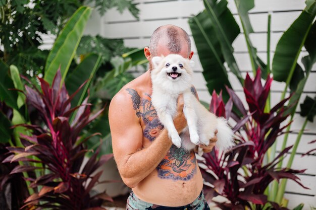 Il giovane uomo felice tatuato brutale barbuto bello tiene lo spitz pomeranian che gioca con l'animale domestico adorabile