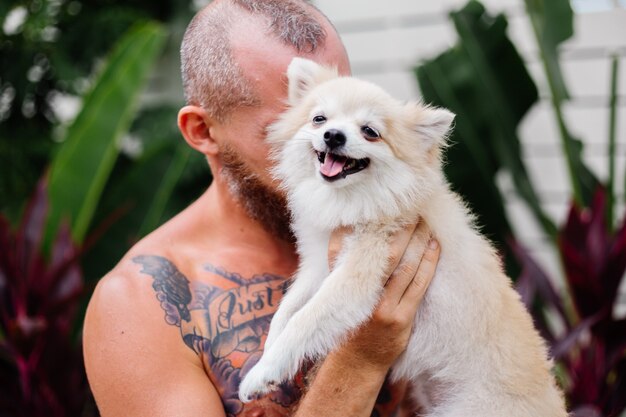 Il giovane uomo felice tatuato brutale barbuto bello tiene lo spitz pomeranian che gioca con l'animale domestico adorabile