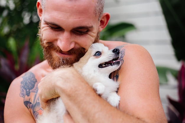 Il giovane uomo felice tatuato brutale barbuto bello tiene lo spitz pomeranian che gioca con l'animale domestico adorabile