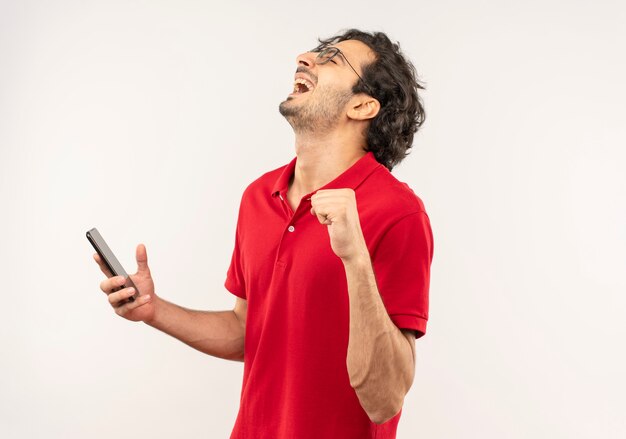 Il giovane uomo gioioso in camicia rossa con vetri ottici tiene il telefono e solleva il pugno isolato sul muro bianco