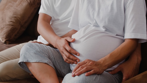 Il giovane uomo incinto asiatico delle coppie tocca la sua pancia della moglie che parla con suo figlio. Mamma e papà sentirsi felici sorridenti pacifici mentre abbi cura del bambino, gravidanza sdraiata sul divano nel salotto di casa.