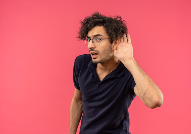 Il giovane uomo infastidito in camicia nera con vetri ottici prova a sentire con la mano a coppa all'orecchio isolato sulla parete rosa