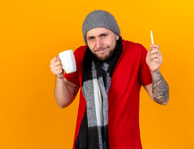 Il giovane uomo malato caucasico infastidito che indossa il cappello e la sciarpa di inverno tiene la tazza ed il termometro isolati sulla parete arancione con lo spazio della copia