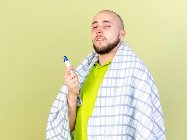 Il giovane uomo malato caucasico pallido avvolto in un plaid tiene il termometro su verde oliva