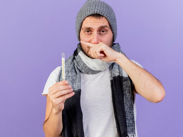 Il giovane uomo malato dispiaciuto che porta il cappello di inverno con il termometro della tenuta della sciarpa che pulisce il naso con la mano isolata su fondo viola