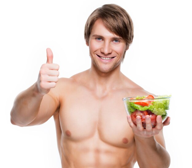 Il giovane uomo muscoloso felice che tiene un'insalata e mostra i pollici aumenta il segno sopra il muro bianco.