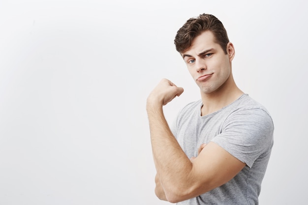 Il giovane uomo muscoloso positivo indossa una maglietta grigia, mostra i bicipiti dopo l'allenamento in palestra, dimostrando quanto sia forte. Deridere, fare facce maschili si vanta della sua forza, dimostrando il suo braccio forte