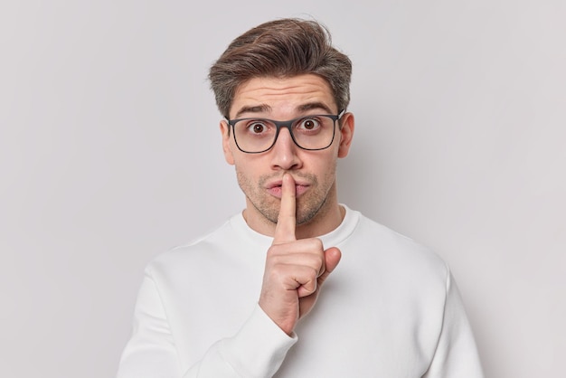 Il giovane uomo segreto con la barba lunga fa il gesto del silenzio preses indxx dito sulle labbra dice che indossa occhiali e un maglione casual chiede di essere tranquillo isolato su sfondo bianco. Concetto di linguaggio del corpo