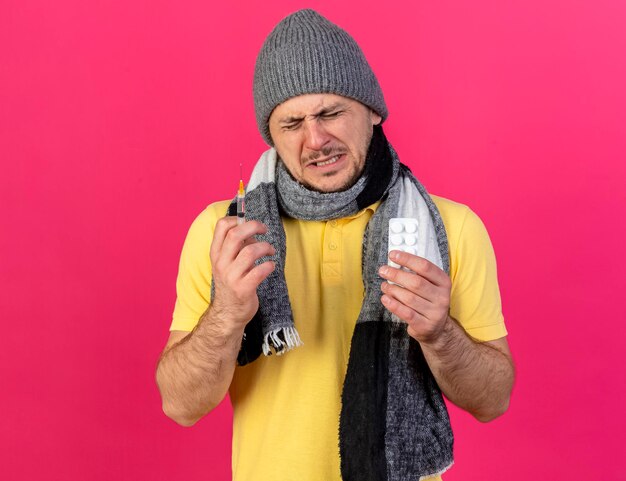 Il giovane uomo slavo malato biondo dispiaciuto che indossa cappello e sciarpa di inverno tiene la siringa