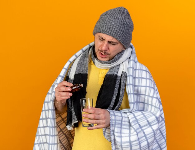 Il giovane uomo slavo malato biondo dispiaciuto che indossa il cappello e la sciarpa di inverno avvolti in plaid tiene il bicchiere d'acqua e la medicina in una bottiglia di vetro isolata sulla parete arancione con lo spazio della copia