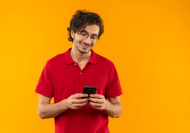Il giovane uomo sorridente in camicia rossa con vetri ottici tiene e guarda il telefono isolato sulla parete arancione