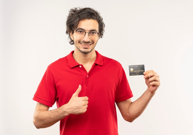 Il giovane uomo sorridente in camicia rossa con vetri ottici tiene la carta di credito e il pollice in alto isolato sul muro bianco
