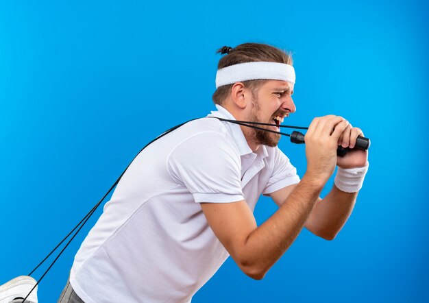 Il giovane uomo sportivo bello dispiaciuto che indossa la fascia e i braccialetti in piedi nella vista di profilo che tiene e che tira la corda di salto isolata sullo spazio blu