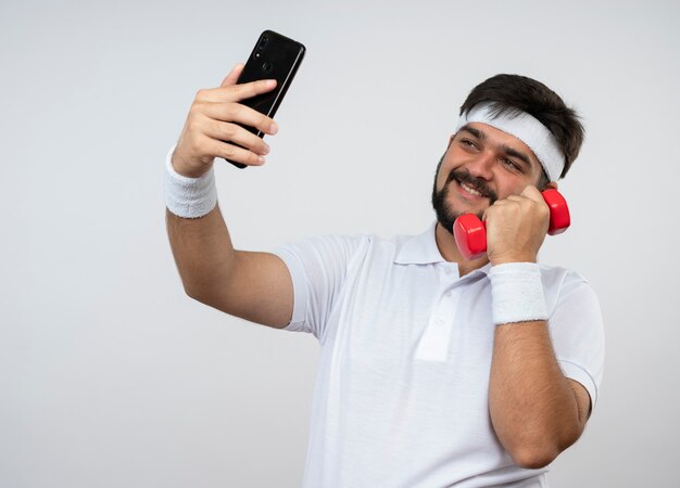 Il giovane uomo sportivo sorridente che indossa la fascia e il polsino che tiene il manubrio prende un selfie isolato sulla parete bianca