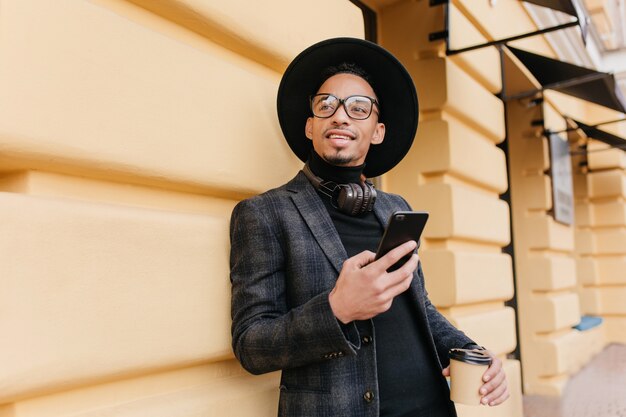 Il giovane vago indossa un vestito nero alla moda che distoglie lo sguardo mentre levandosi in piedi sulla strada con la tazza di tè. Ragazzo africano occupato in attesa di qualcuno e che tiene smartphone e caffè.