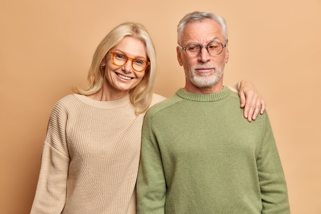 Il marito e la moglie anziani posano per il sorriso dell'abbraccio del ritratto di famiglia vestito positivamente con i ponticelli degli occhiali che si levano in piedi contro il muro marrone dello studio