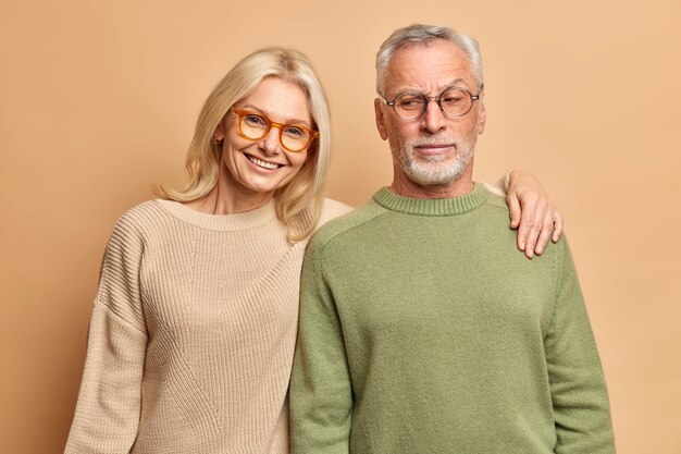 Il marito e la moglie anziani posano per il sorriso dell'abbraccio del ritratto di famiglia vestito positivamente con i ponticelli degli occhiali che si levano in piedi contro il muro marrone dello studio