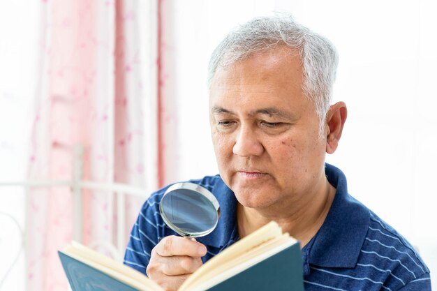 Il maschio anziano felice dell'uomo anziano si diverte a leggere il libro usando la lente d'ingrandimento