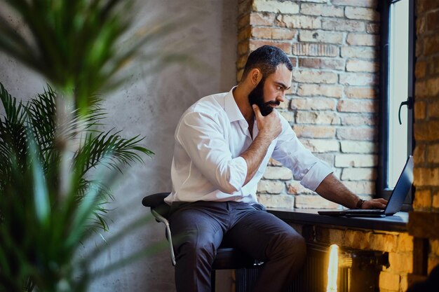 Il maschio barbuto alla moda lavora con un laptop in una stanza con interni soppalcati.