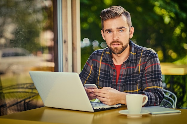 Il maschio barbuto bello beve il caffè mentre usa un computer portatile in un caffè.