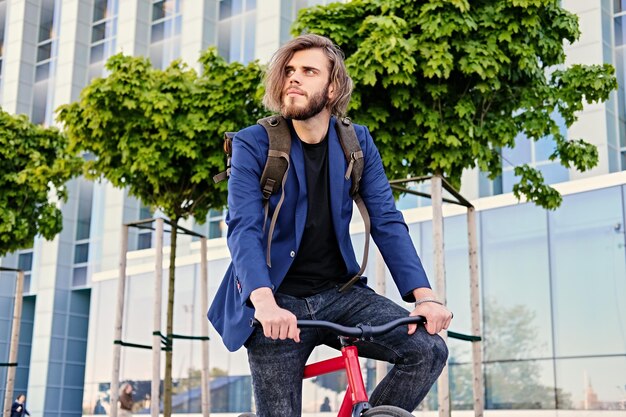 Il maschio barbuto dei pantaloni a vita bassa con lo zaino si siede sulla bicicletta fissa rossa in un parco.