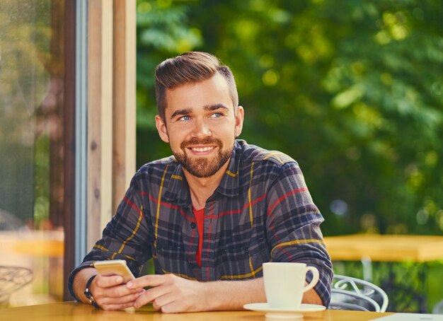 Il maschio barbuto sorridente beve il caffè e utilizza lo smartphone in un bar.