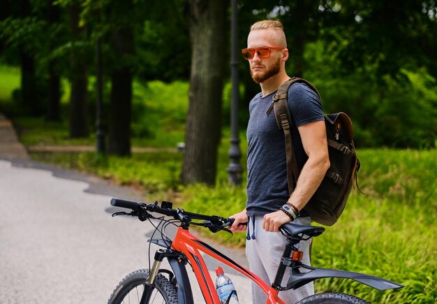 Il maschio barbuto sportivo si siede su una bicicletta di montagna rossa all'aperto.