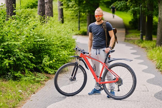 Il maschio barbuto sportivo si siede su una bicicletta di montagna rossa all'aperto.