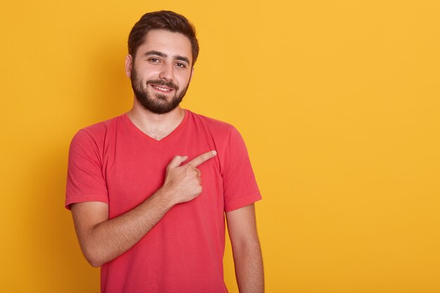 Il maschio con la barba lunga bello orizzontale, vestito in maglietta rossa casuale, indicando da parte il dito indice, mostra lo spazio della copia per la vostra pubblicità o il testo di promozione.