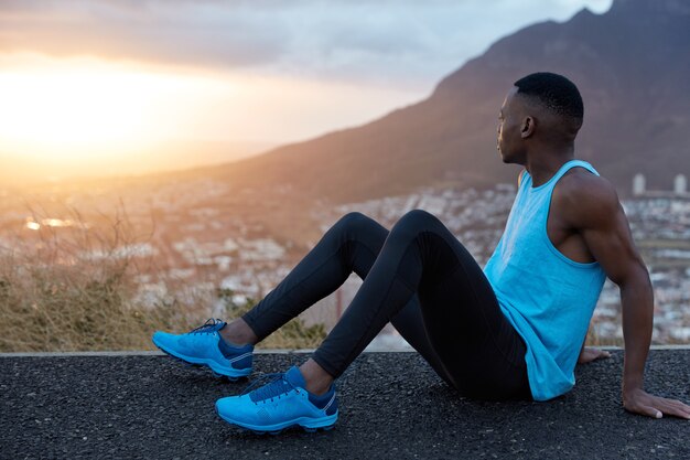 Il maschio fitness si siede di lato, ha la pelle nera, le mani muscolose, vestito con abbigliamento sportivo, guarda attentamente all'alba, posa sulle montagne, si prende una pausa dopo una corsa intensa. Sport, natura