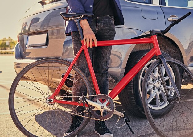 Il maschio hipster barbuto con i capelli lunghi tiene una bicicletta a velocità singola vicino all'auto.