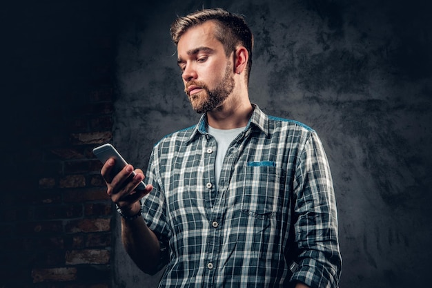 Il maschio hipster barbuto in una maglietta in pile tiene lo smartphone su sfondo grigio.