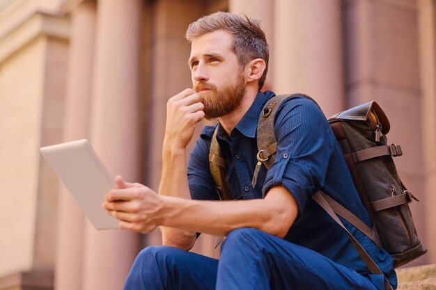 Il maschio viaggiatore casual barbuto si siede su un gradino e utilizza un tablet PC.