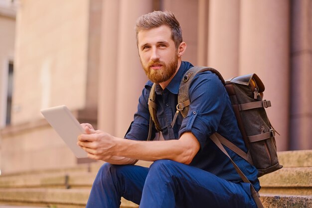 Il maschio viaggiatore casual barbuto si siede su un gradino e utilizza un tablet PC.