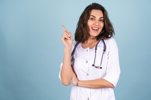 Il medico della giovane bella donna in un camice da laboratorio su sfondo blu ha improvvisamente avuto un'idea, punta il dito verso l'alto