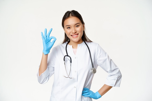 Il medico femminile asiatico sorridente, l'infermiera in guanti di gomma e l'uniforme medica mostrano il segno ok, approva e gli piace smth buono, in piedi su sfondo bianco