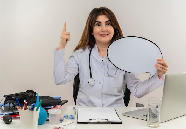 Il medico femminile di mezza età soddisfatto che indossa la veste medica con lo stetoscopio che si siede allo scrittorio lavora sul computer portatile con gli strumenti medici che tengono la bolla di chiacchierata e indica in su sul muro bianco isolato