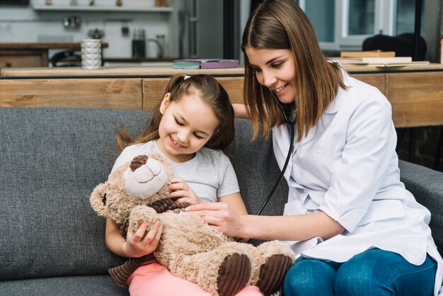 Il medico femminile sorridente che esamina l&#39;orsacchiotto tiene dalla ragazza felice