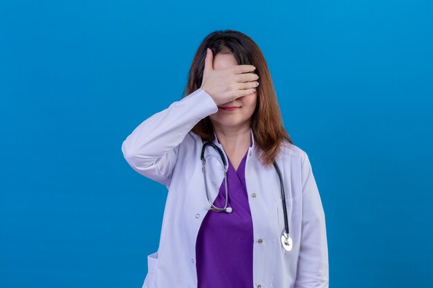 Il medico invecchiato centrale che indossa le camice bianche e con la copertura dello stetoscopio osserva con la mano che sorride sopra la parete blu