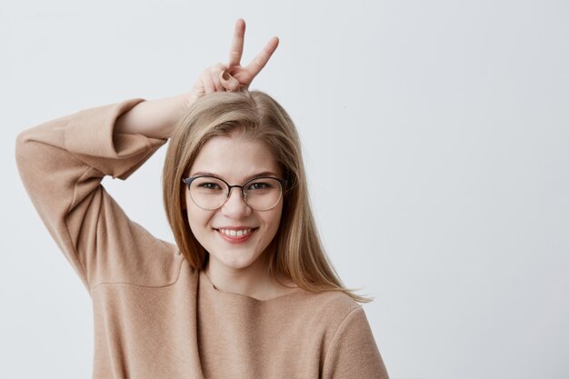 Il modello femminile adorabile sveglio in occhiali sembra allegro, gesti con le dita, ha espressione felice, posa con lo spazio della copia. Ragazza bionda graziosa divertendosi