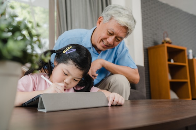Il nonno asiatico insegna alla nipote che disegna e che fa i compiti a casa. Il cinese senior, nonno felice si rilassa con la ragazza che si trova sul sofà nel concetto del salone a casa.