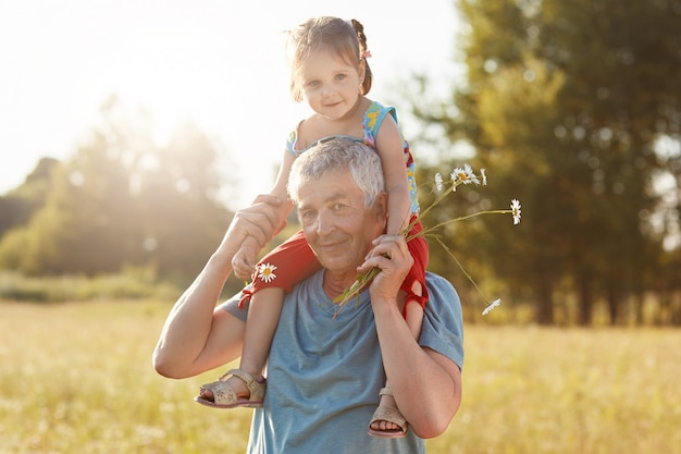 Il nonno e la nipote felici si divertono insieme all'aperto. Maschio dai capelli grigi dare cavalluccio per bambino piccolo