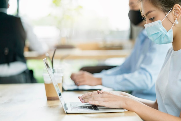 Il nuovo concetto di stile di vita normale, gli uomini d'affari che indossano la maschera si siedono con l'allontanamento sociale lavorando in aree pubbliche o in uno spazio di lavoro condiviso con laptop e smartphone